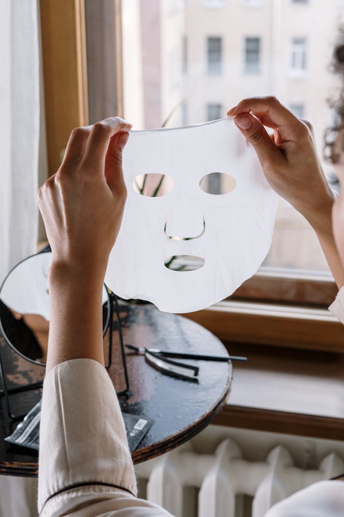 A woman holds a sheet mask in a cozy home setting, by a window.